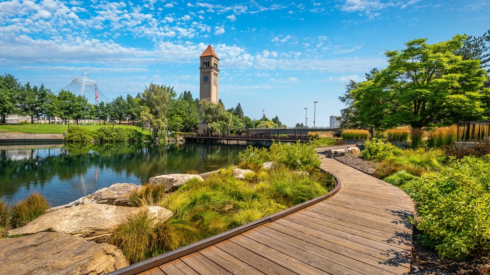 A scenic park pathway leading towards a historic clock tower with a serene pond in the foreground.