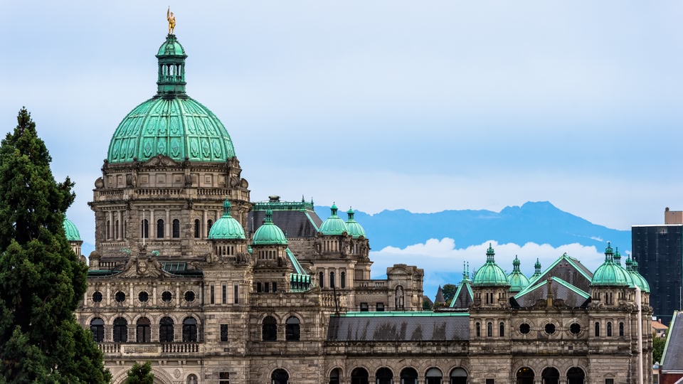 The British Columbia Parliament Buildings in Victoria, featuring a grand, domed structure with green copper roofing and intricate stonework, set against a backdrop of mountains.