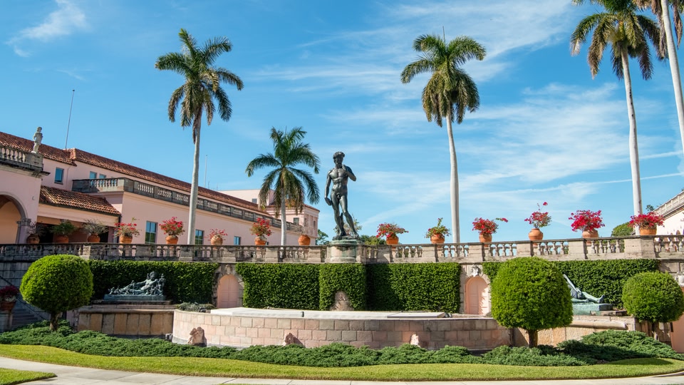 A landscaped courtyard with a central statue, surrounded by hedges and palm trees, and buildings in the background.